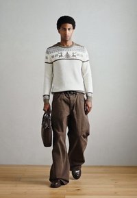 Hrubý biely pletený sveter so vzorom sobov, v kombinácii s voľnými hnedými nohavicami. Drží hnedú koženú tašku; stojí na drevenej podlahe.