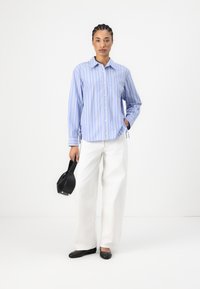 Striped blue and white button-up shirt paired with white wide-leg pants. Model holds a black handbag and wears flat black shoes.