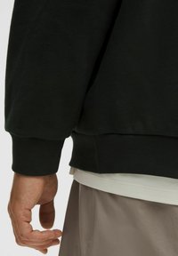 Sort sweatshirt med ribbede manchetter og kant, der har en subtil logo-detalje på ryggen, lagdelt over en lys skjorte.
