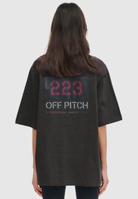 Sort oversized t-shirt med iøjnefaldende grafik på ryggen: "223 OFF PITCH" med koordinater, i kontrasterende rød og grå tekst.