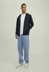 Homem em pé, vestido com um hoodie azul escuro, t-shirt branca, jeans azul claro e ténis brancos, em fundo neutro.