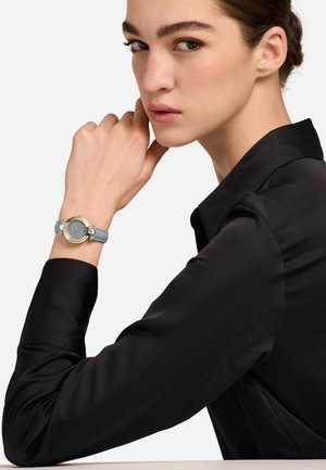 Reloj redondo con marco dorado, correa de cuero gris claro y esfera azul texturizada. El modelo, vestido con una camisa negra, descansa la barbilla en la mano.
