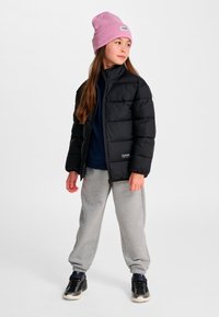 Ung person iført en lyserød beanie, sort pufferjakke, grå joggingbukser og sorte sneakers, stående og kiggende til siden mod en hvid baggrund.