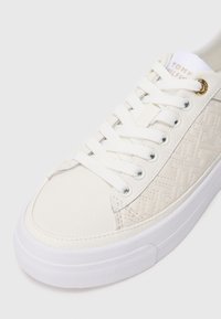 White sneaker with a textured leather upper, featuring a geometric pattern and white laces. Metal eyelet accents and rubber sole.