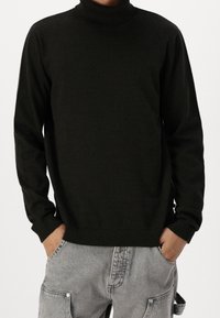 Osoba w czarnym swetrze z golfem i jasnoszarych dżinsach, z rękami w kieszeniach, stojąca na jednolitym tle.