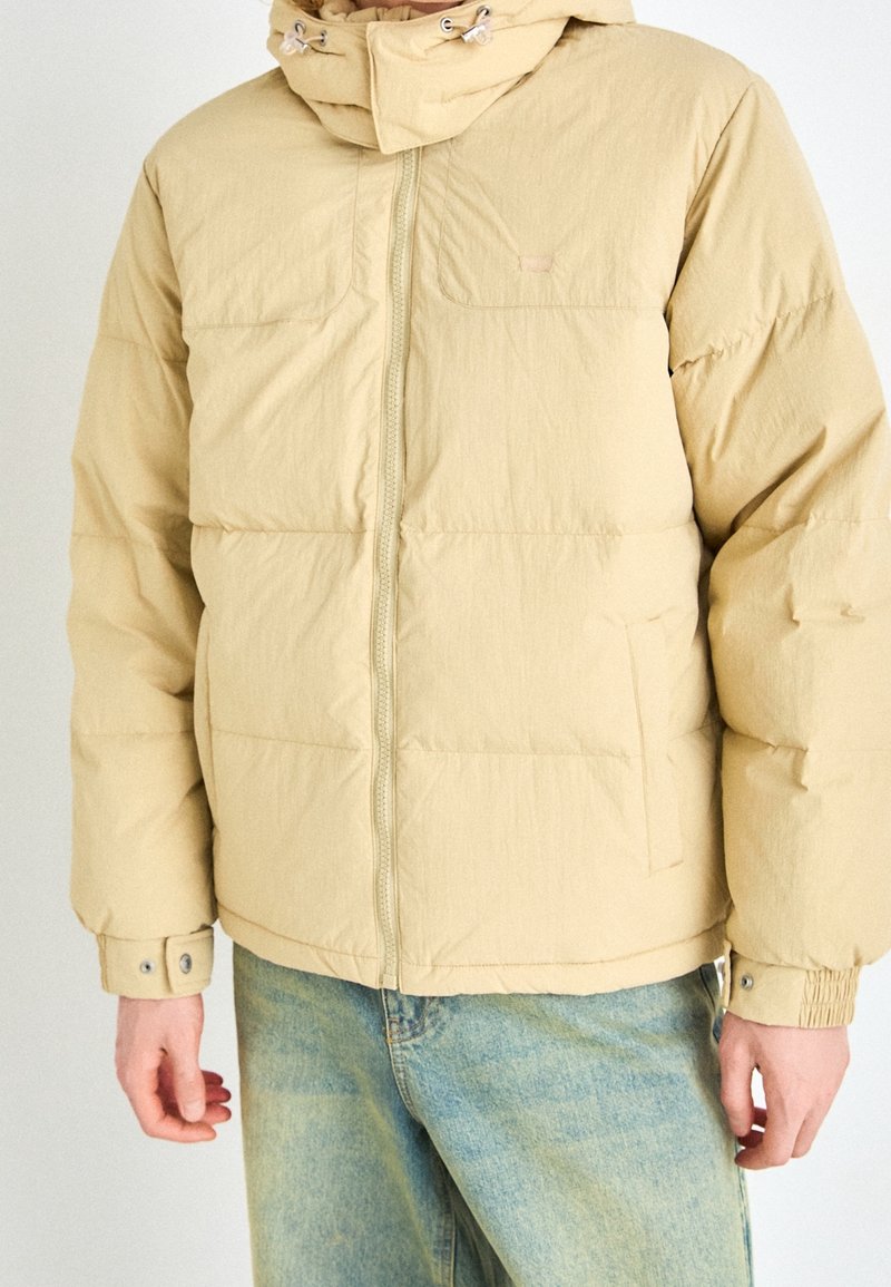 Personne portant une doudoune beige avec fermeture éclair et capuche, associée à un jean en denim bleu clair, se tenant devant un fond uni.