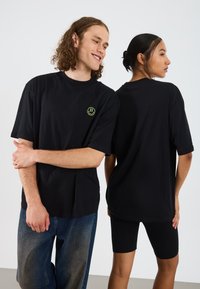 Sort oversized T-shirt lavet af bomuld, med et lille grønt smiley ansigt logo på brystet. Båret med denim og sorte leggings.