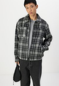 Gray plaid jacket with a collared neckline, zip front, two chest pockets, and long sleeves, paired with dark gray pants and a black bag.