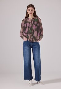 Junge Frau steht mit einem mehrfarbigen, pink- und grünmusterierten Blusen, blauen geraden Jeans und weißen Turnschuhen da, mit den Händen in den Taschen.