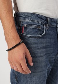 Dunkelblaue Jeans aus Denim mit einem roten Etikett, kombiniert mit einem schwarzen Kettenarmband an einer Hand, die auf der Hüfte ruht, und damit einen lässigen Look zeigt.