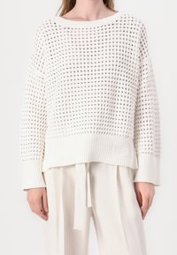 Woman wearing a white open-knit sweater with long sleeves and ribbed cuffs paired with loose white pants with a fabric belt.