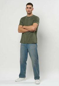 Olivengrønn bomulls-T-skjorte med rund hals, kombinert med lyseblå denimjeans. Modellen står med armene i kors og har på seg hvite joggesko.