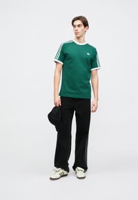 Πράσινο βαμβακερό μπλουζάκι με λευκό γιακά και τρεις λευκές ρίγες στους ώμους, συνδυασμένο με φαρδιά μαύρα παντελόνια και κρεμ αθλητικά παπούτσια. Μαύρο καπέλο κρατημένο στο χέρι.