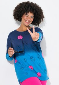 Camicia a maniche lunghe blu ombre con stampe di faccine sorridenti in rosa e nero, abbinata a pantaloni rosa brillante. Modella mentre tiene occhiali da sole.