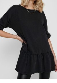 Kobieta w luźnej, czarnej sukience z długim rękawem i falbanym dołem, z warstwami złotych naszyjników i czarnymi legginsami, pozująca z ręką na biodrze.