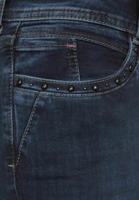 Mörkblå denimjeans med svarta nitar längs framfickan. Har synlig sömnad och en strukturerad yta. Slim fit-design.