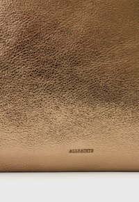 Metallic guld läderpåse med texturerad yta och präglad "ALLSAINTS"-logotyp längst ner. Rektangulär form, slät finish.