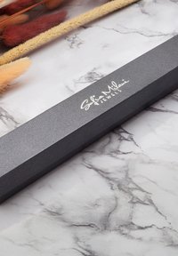 Scatola rettangolare nera con superficie testurizzata, con scritte bianche che dicono "Sofia Milani Jewels", posizionata su uno sfondo a motivo marmorizzato.