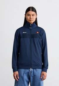 Mornarska zipped jakna z visokim ovratnikom, ki ima črn panel čez prsa, ellesse logo in kontrastni barvni poudarek nad prsmi.