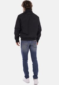 Schwarze Bomberjacke mit hohem Kragen, Rippbündchen und Saum, getragen über blauen Denim-Jeans. Die Rückansicht zeigt ein minimalistisches Design und eine glatte Textur.