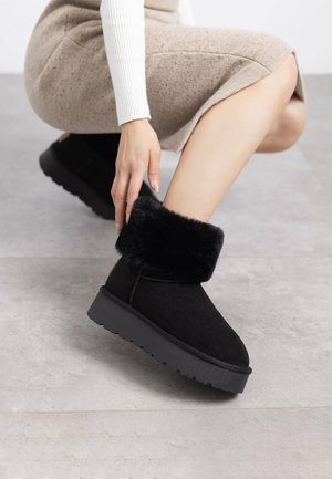 Stivali alla caviglia neri in suede, con un morbido risvolto in pelliccia sintetica e una suola spessa e testurizzata. Il design è minimalista, senza hardware visibili.