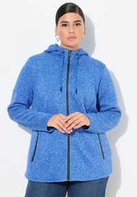 Blå huvjacka med en texturerad yta. Har en hel dragkedja och två sidofickor med svarta dragkedjor. Mjuk, avslappnad design.