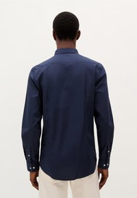 Cămașă albastră închis cu mâneci lungi, cu un design ajustat, guler cu nasturi și manșete cu nasturi albi. Textură netedă și formă adaptată.