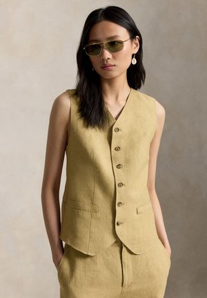 Nő, bézs színű ujjatlan gombos mellényt és hozzáillő nadrágot visel, zöldes színű napszemüveggel és kagylófülbevalóval, semleges háttér előtt áll.