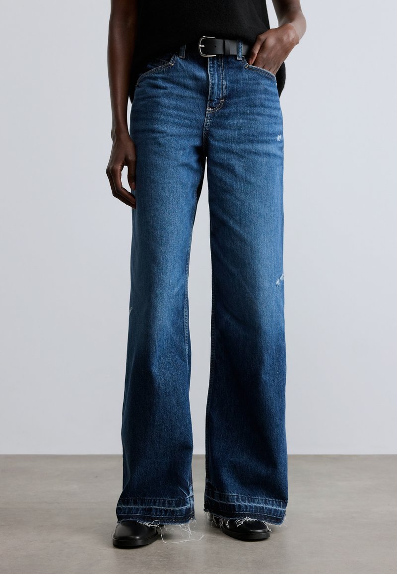 Széles szárú kék farmernadrág foszlott szegéllyel, közepes derékmagassággal és enyhe kopott hatással. Fekete övvel és felsővel párosítva.