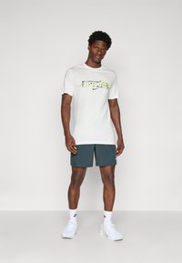 Camiseta blanca con texto "JUST DO IT" en patrón verde, combinada con pantalones cortos verde oscuro. El modelo lleva zapatillas blancas y calcetines.