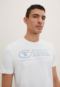 Hvid bomulds T-shirt med blå trykt tekst og logo, korte ærmer, rund halsudskæring og afslappet pasform.