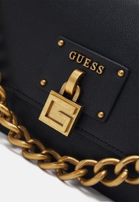 Mustakalloinen musta nahkalaukku, jossa on kultaketju, kultalukkosomiste ja kohokuvioitu "GUESS"-logo suorakaiteen muotoisessa tarrassa.