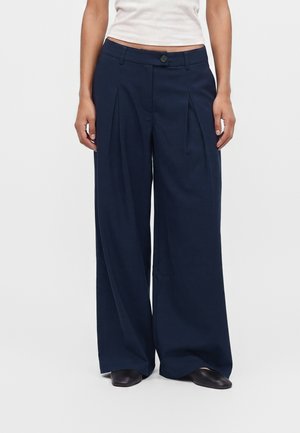 Woman wearing dark blue wide-leg trousers with front pleats, a button and zipper, paired with black shoes and a white top.