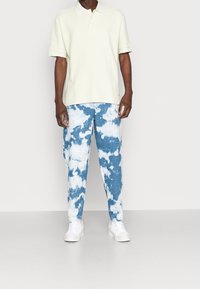 Světle žluté bavlněné polo tričko s krátkými rukávy, v kombinaci s modrými džínovými kalhotami s batikovaným vzorem a bílými obláčky, doplněné bílými teniskami.