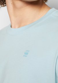Světle modré bavlněné tričko s kulatým výstřihem, které na hrudi má malé, decentní logo tištěné v ladící barvě. Hladká textura.