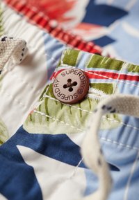 Tkanina w kwiatowy wzór, głównie niebieska i zielona, z brązowym guzikiem z wytłoczeniem "SUPERDRY". Teksturowana powierzchnia z czerwonymi akcentami.