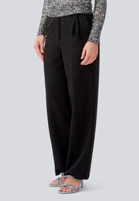 Black wide-leg trousers made of smooth fabric, featuring a tied waistband. Paired with strappy light blue heeled sandals.