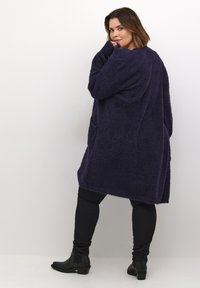 Lang lilla fluffy cardigan med løs pasform, sidelommer og dropped shoulders, parret med sorte skinny bukser og ankelstøvler.