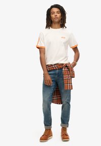 Hvid t-shirt med orange kant og logo, blå jeans, brune læderstøvler, og en rød ternet skjorte bundet om livet.