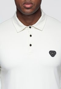 Weißes Polohemd mit einem Kragen, drei schwarzen Knöpfen und einem dreieckigen schwarzen Logopatch auf der linken Brustseite. Glatte Textur.