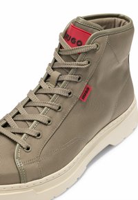 Beige lærred høj-top sneakers med rund tå, tekstureret overflade og kontrasterende rød logo på tungen og siden, udstyret med snørebånd.