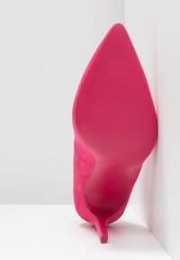 Escarpin en suède rose avec un bout pointu et une semelle lisse et texturée. Le talon est fin et mesure environ quatre pouces de haut.