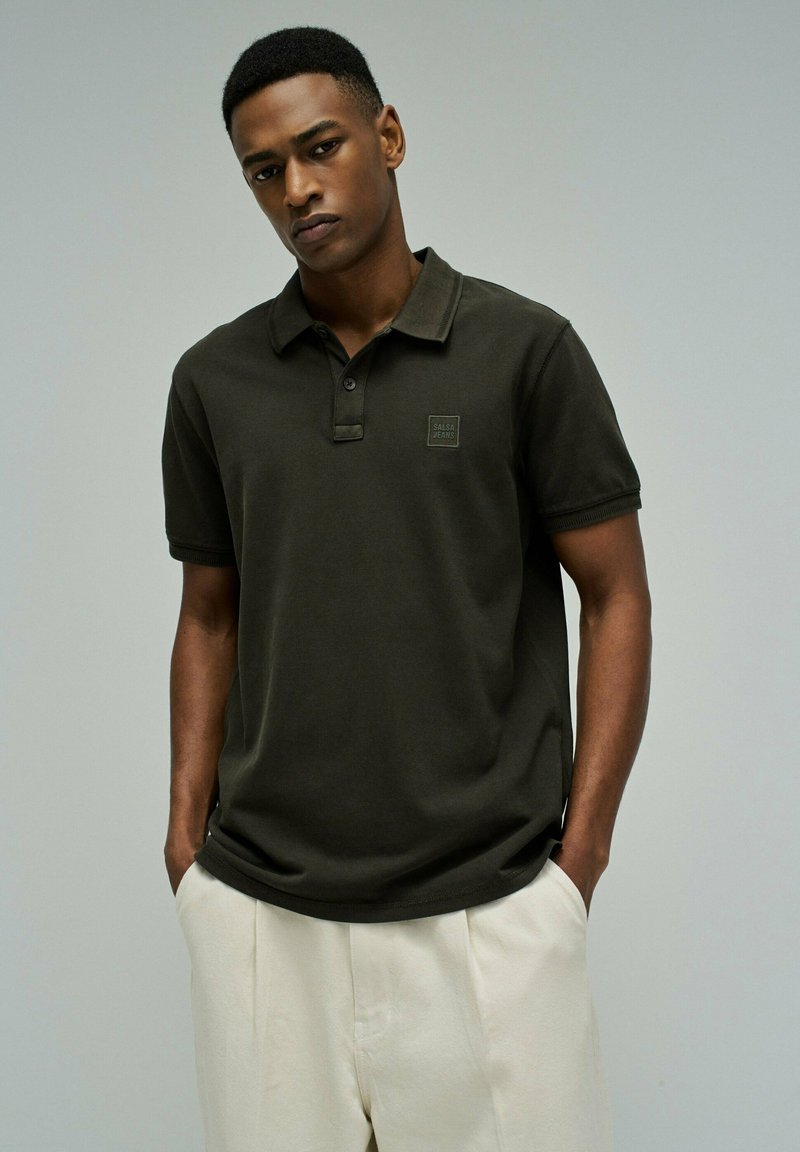 Junger Mann trägt ein dunkelgrünes Poloshirt mit dem "Salsa Jeans"-Logo, steht mit den Händen in den Taschen vor einem schlichten grauen Hintergrund.