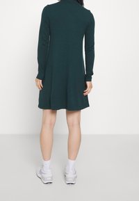 Robe à manches longues en tissu maille vert foncé, forme évasée, encolure arrondie, longueur aux genoux, associée à des baskets blanches et des chaussettes montantes.
