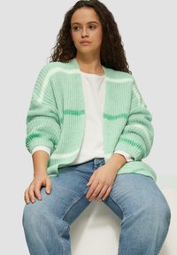 Minzgrüner, offener Strickcardigan mit weißen und dunkler grünen Streifen, oversized Ärmeln und lockerer Passform, getragen über einem weißen Hemd und blauen Jeans.