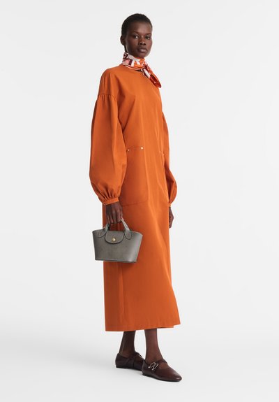 Frau in einem langärmeligen orangefarbenen Kleid, mit einem gemusterten Halstuch, braunen Ballerinas und einer kleinen grauen Handtasche vor einem schlichten Hintergrund.