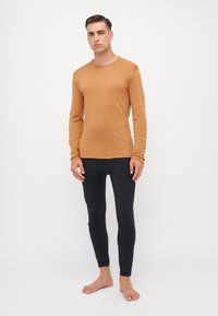 Chemise à manches longues, col rond, couleur rouille, en tissu doux, associée à des leggings noirs ajustés avec une texture lisse.