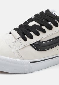 Adidași din suede alb cu șireturi negre și un accent din piele neagră. Prezintă cusături gri și o talpă din cauciuc alb texturat.