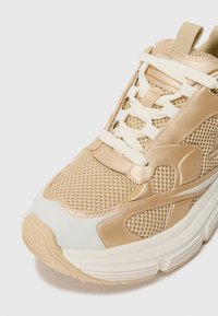 Sneaker a rete beige e crema con suola spessa, lacci bianchi e linguetta con logo, mostrato in primo piano su uno sfondo neutro.