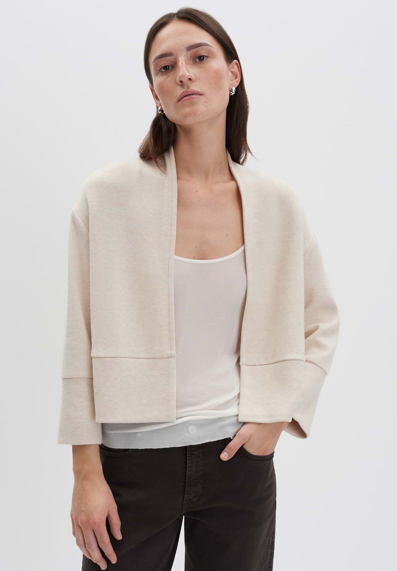 Beige oversized Cardigan aus weichem Material, cropped Design mit weiten Ärmeln, minimalistischer Silhouette und ohne Verschlüsse.